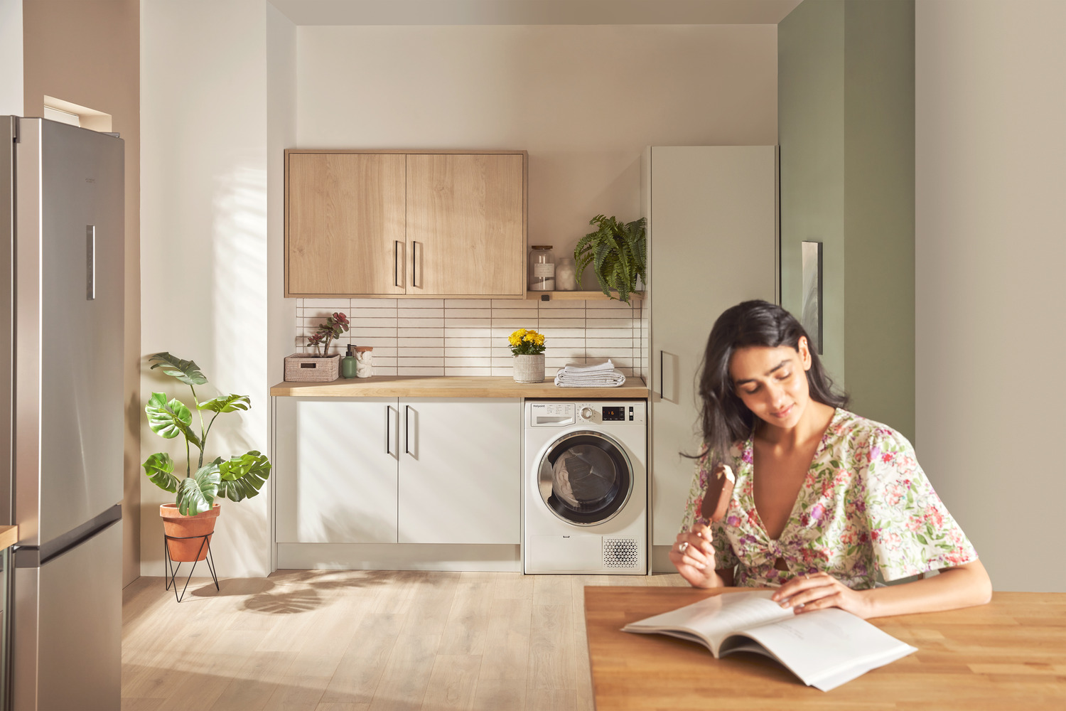 A lady wearing a patterned shirt holding an ice cream cone sitting in front of a bright laundry room with white stacked washing machine, wood-toned cabinets, and potted plants, creating a warm and inviting atmosphere.