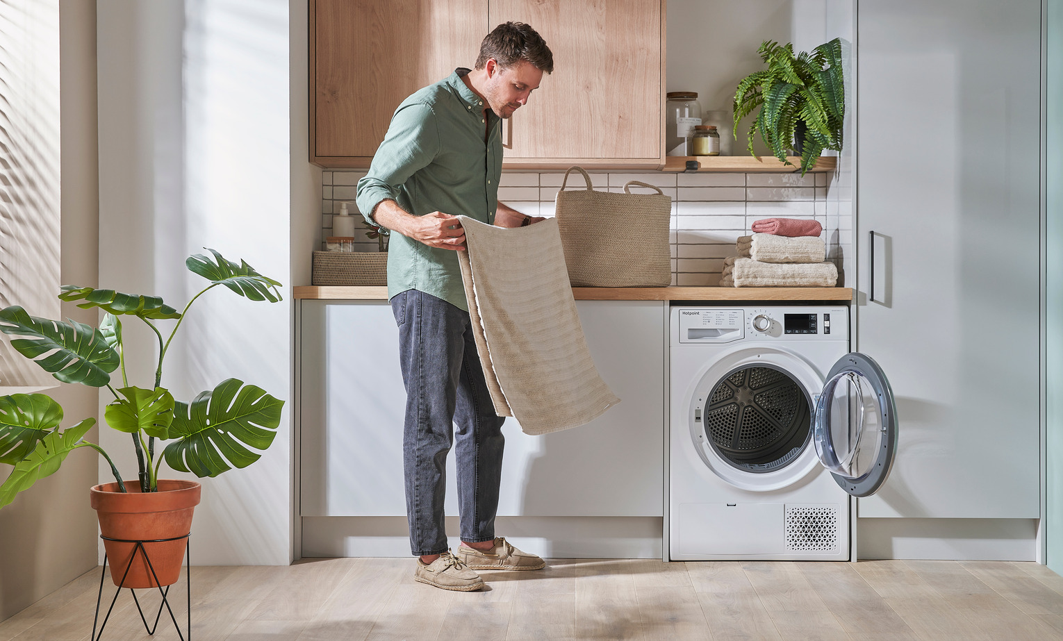 A man stands in front of an open dryer holding a towel in a laundry room with white cabinets, wooden countertops, folded towels, and a potted plant by the window.