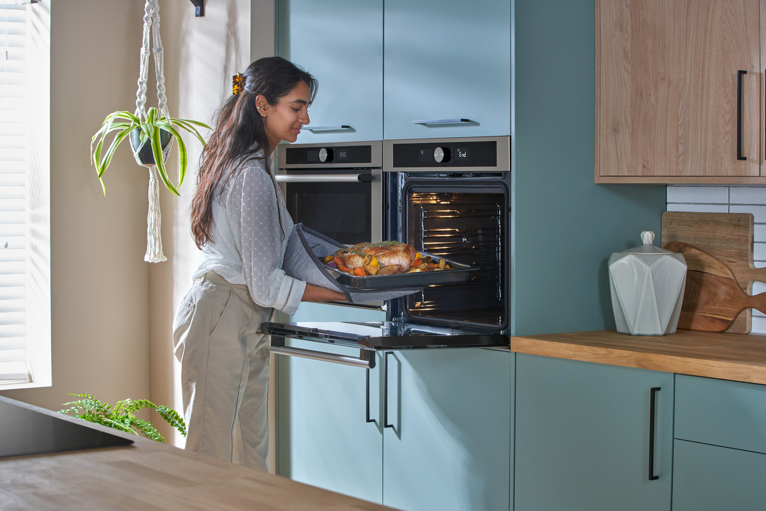 A woman is removing a tray with a cooked chicken and vegetables from a built-in oven in a modern kitchen.
