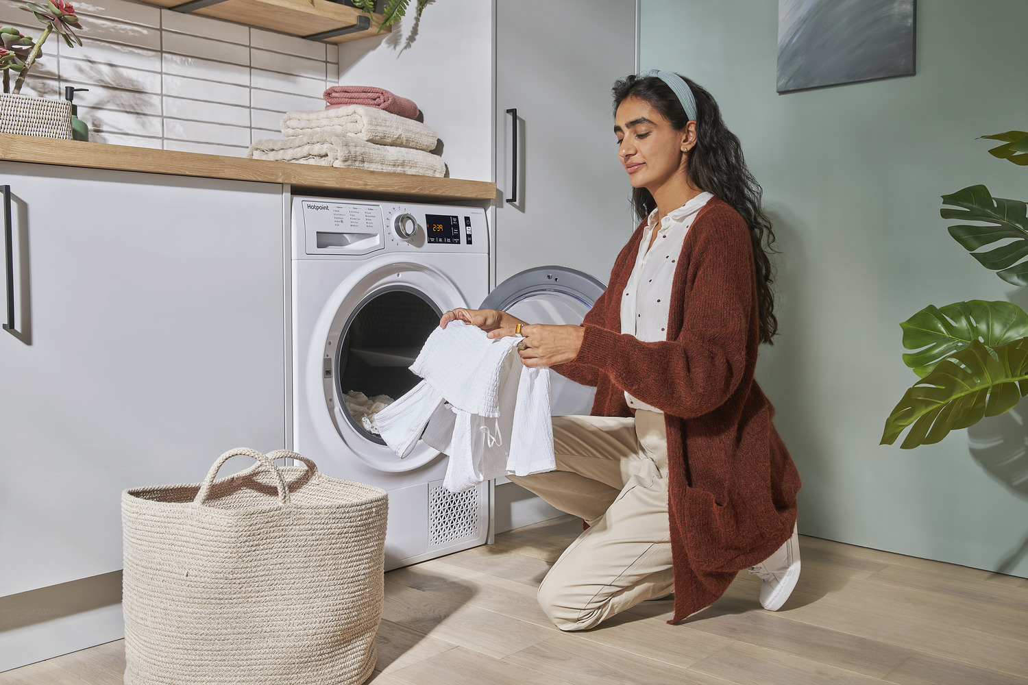 A woman kneels in front of an open dryer, folding white laundry. A laundry basket sits nearby, and the room features white cabinets, wooden countertops, and folded towels.