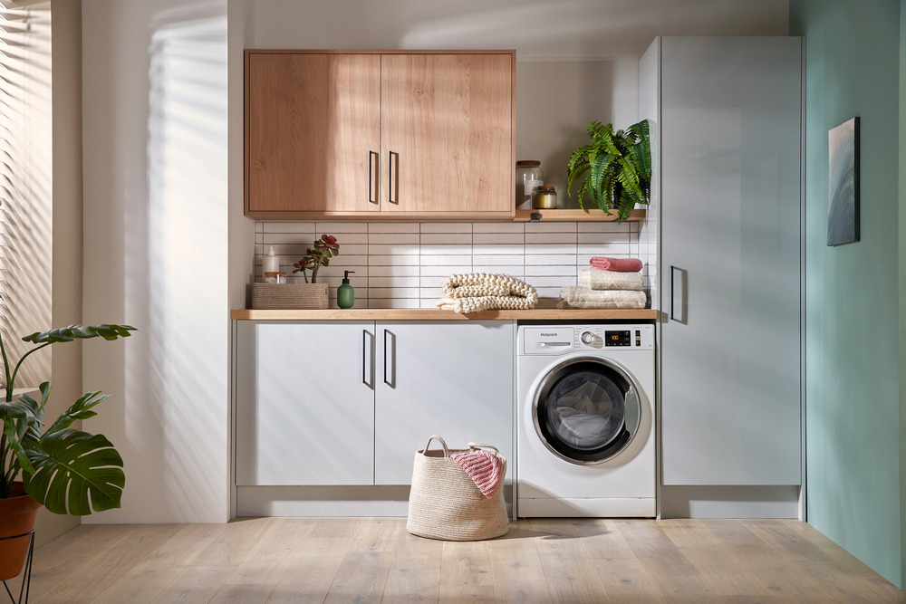A bright laundry room with sleek white cabinets, a warm wood countertop, and a modern white washing machine, accented with green plants and a woven laundry basket on a light wood floor.