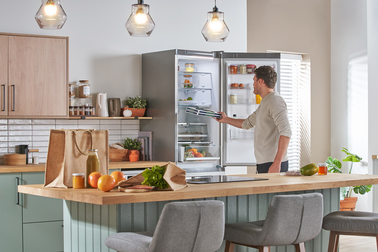 A man placing items into an open refrigerator in a modern kitchen. Fresh produce and reusable grocery bags are on the countertop in the foreground.