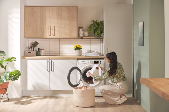 A person is loading laundry into a white washing machine in a bright, modern laundry room with light wood cabinets, white countertops, a white subway tile backsplash, and various plants.