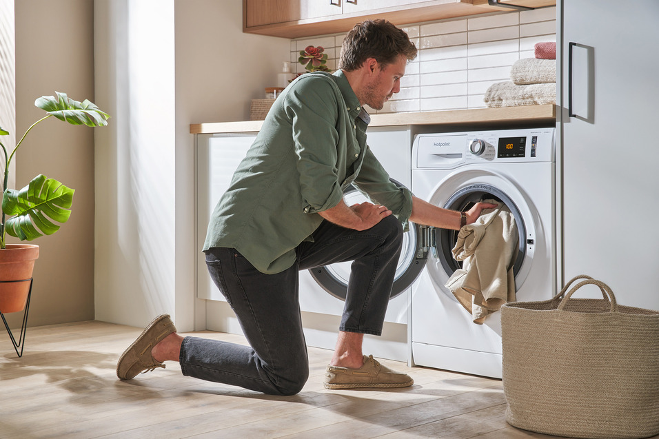 A man in green short crouches in front of a washing machine, transferring laundry from the machine to a rope laundry basket.