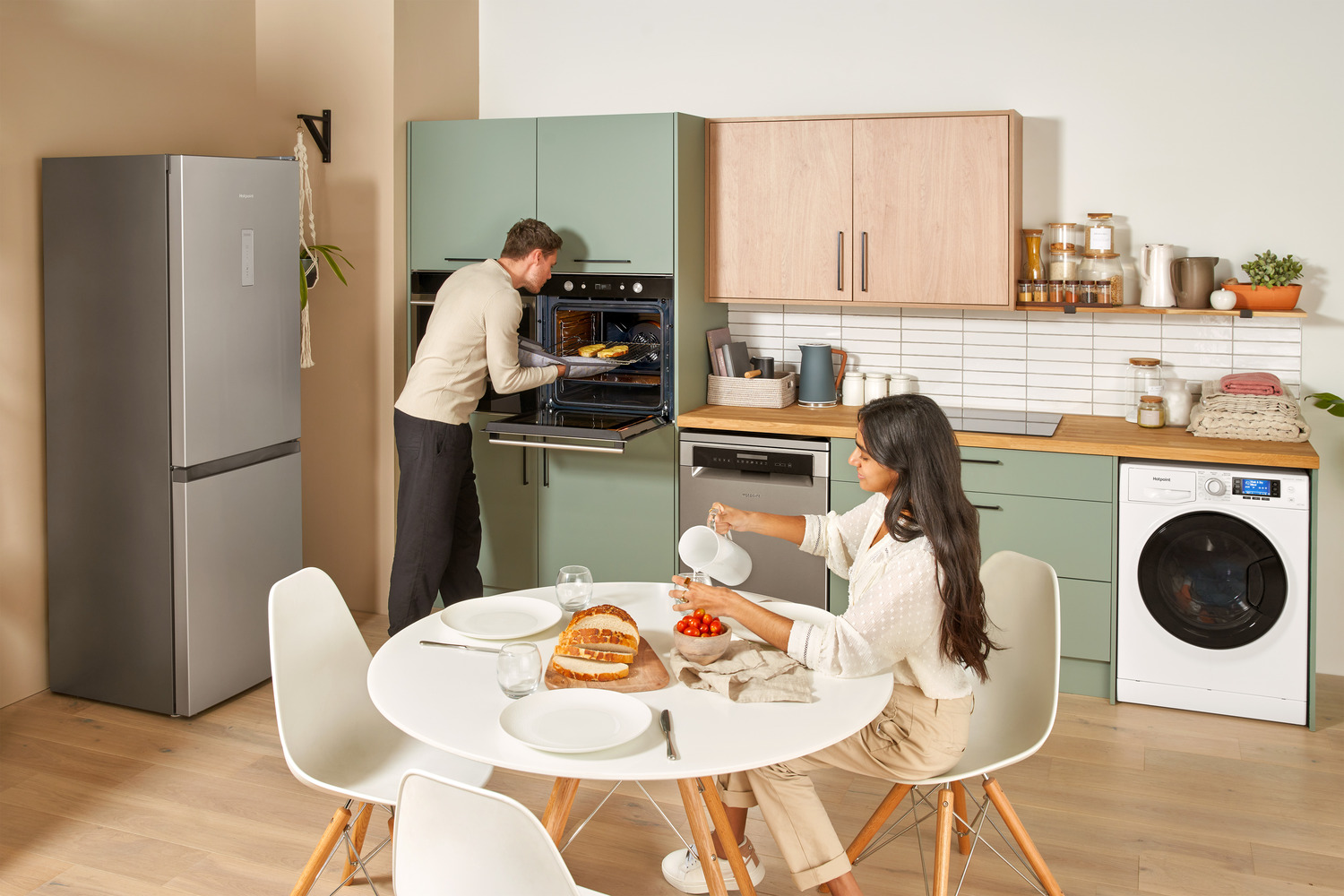 Modern kitchen with sage green cabinets and white appliances, a couple prepares dinner together as the man checks something in the oven while the woman sets a round white dining table with plates, glasses, and a freshly sliced loaf of bread alongside a bowl of cherry tomatoes.