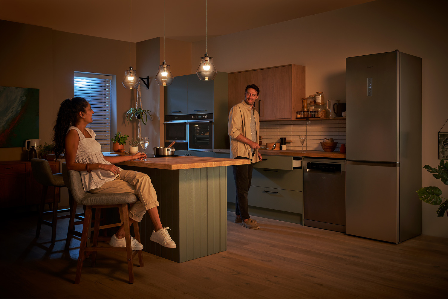 A couple interacts in a dimly lit modern kitchen with pendant lights, where a woman sits at the island counter and a man stands near the oven, surrounded by plants and contemporary appliances in a cozy evening atmosphere.