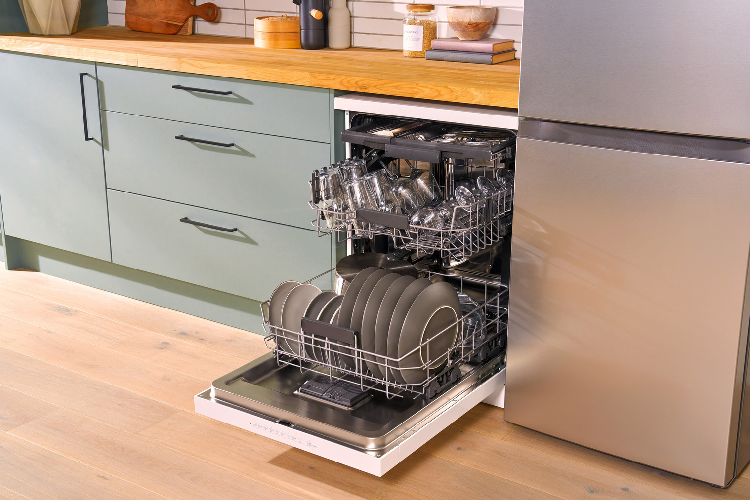 An open dishwasher in a kitchen with light wooden countertops and sage green cabinets. The dishwasher is loaded with neatly arranged plates, bowls, and glassware across three racks.