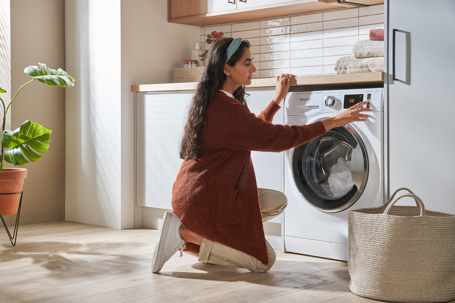 A person in a rust-colored sweater crouches to operate a white washing machine with an open door revealing white laundry inside, next to a large woven laundry basket on a light brown floor