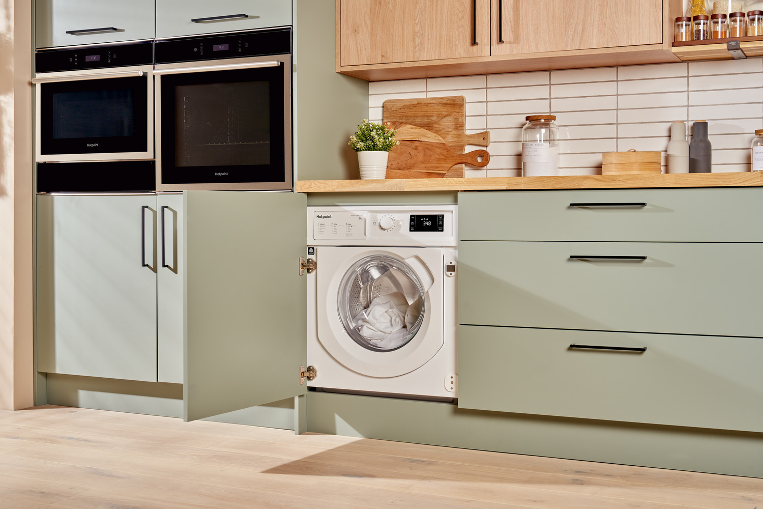 A white Hotpoint washing machine is integrated into a green kitchen cabinet. The washing machine door is open, showing white clothes inside.