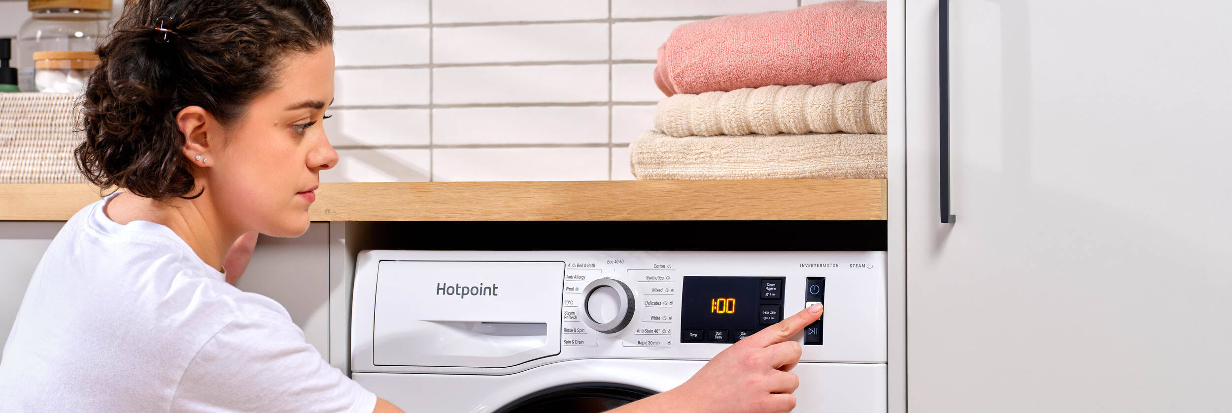 A person is using a Hotpoint washing machine. They are pressing a button on the control panel, which displays "1:00" on a digital screen. Above the machine, there are neatly folded towels in shades of beige and pink on a wooden shelf. The background features white tiled walls.
