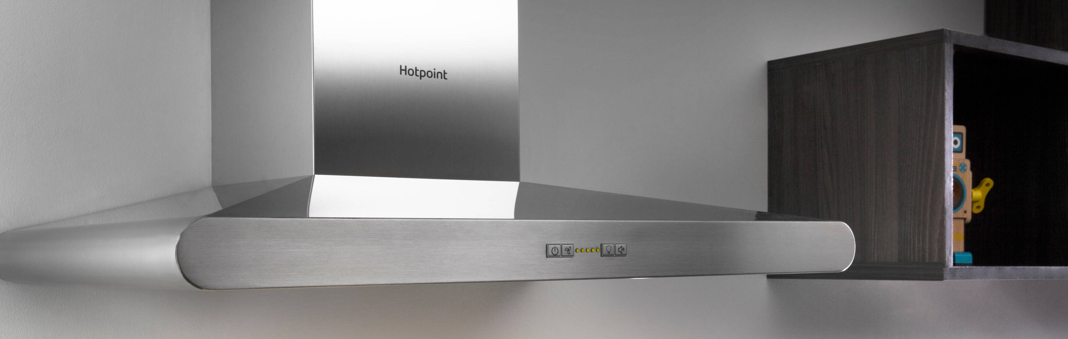 Close-up of a stainless steel range hood with a light turned on, installed beneath wooden cabinets. The brand "Hotpoint" is visible on the control panel.
