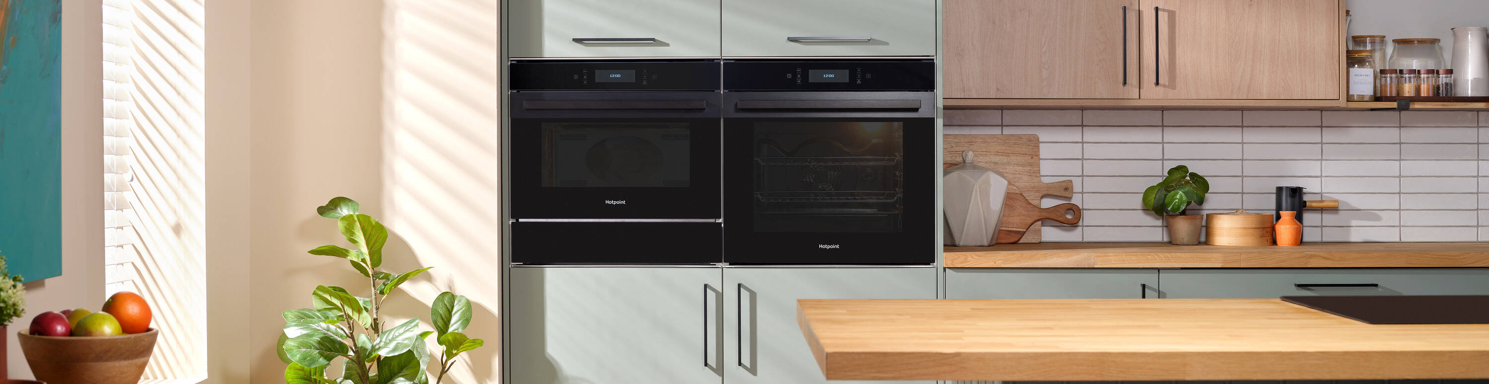 A modern kitchen with two built-in black Hotpoint ovens, light green cabinets, and wood countertops. A hanging plant is near a window, and cutting boards sit on the counter.