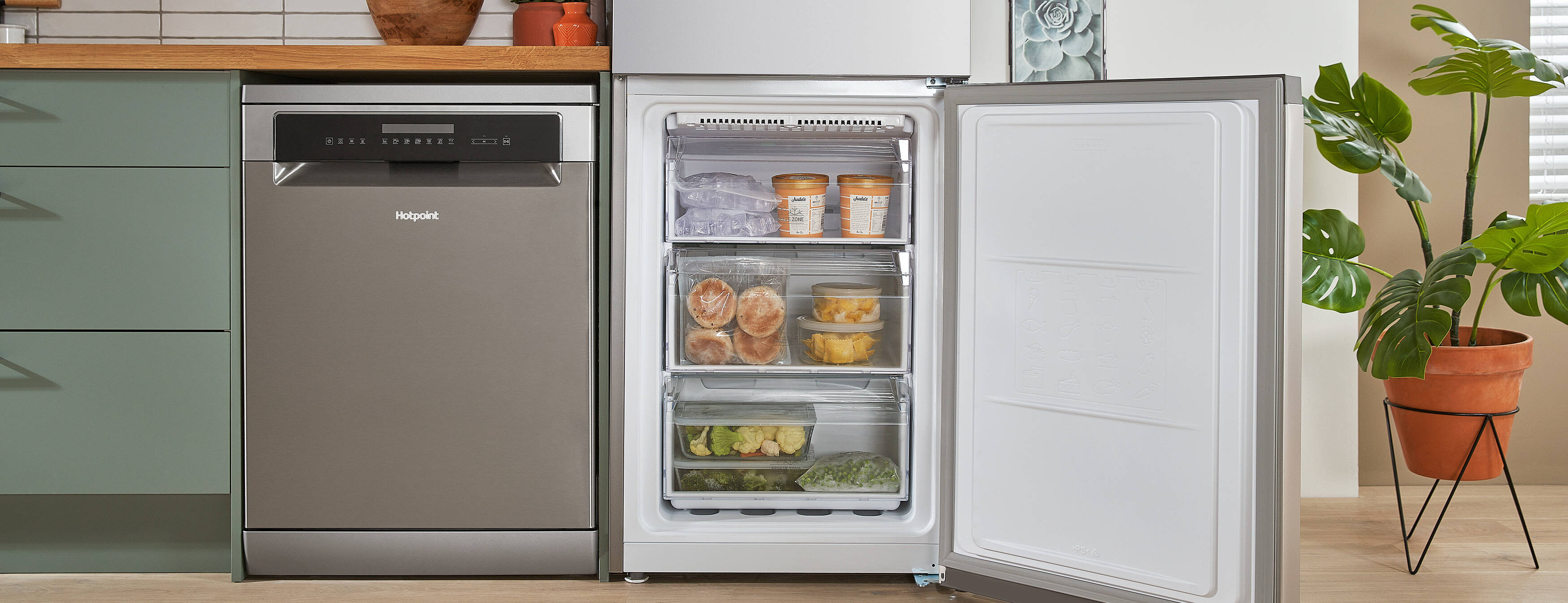 A fridge-freezer with the fridge compartment open, displaying bottles, jars, and fresh produce. It's in a modern kitchen with green cabinets, wood counters, and potted plants.