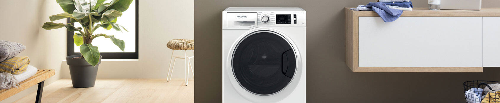 A white Hotpoint washing machine with a black control panel sits in a bright laundry room with a wooden bench and potted plant.