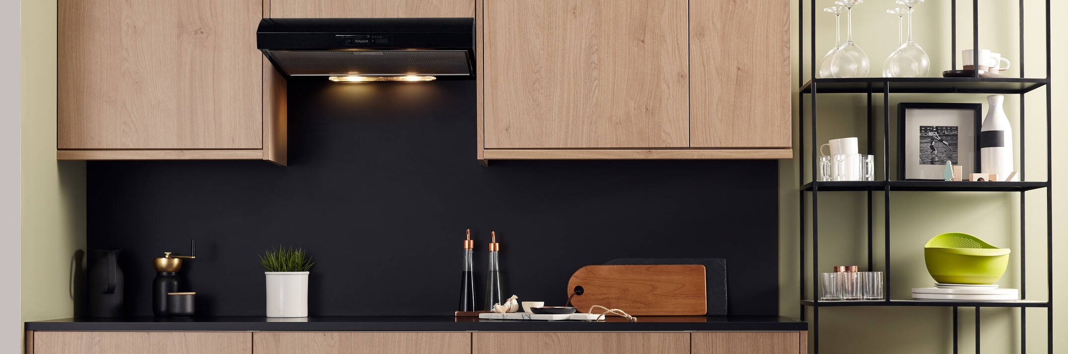 Close-up of a stainless steel range hood with a light turned on, installed beneath wooden cabinets. The brand "Hotpoint" is visible on the control panel.