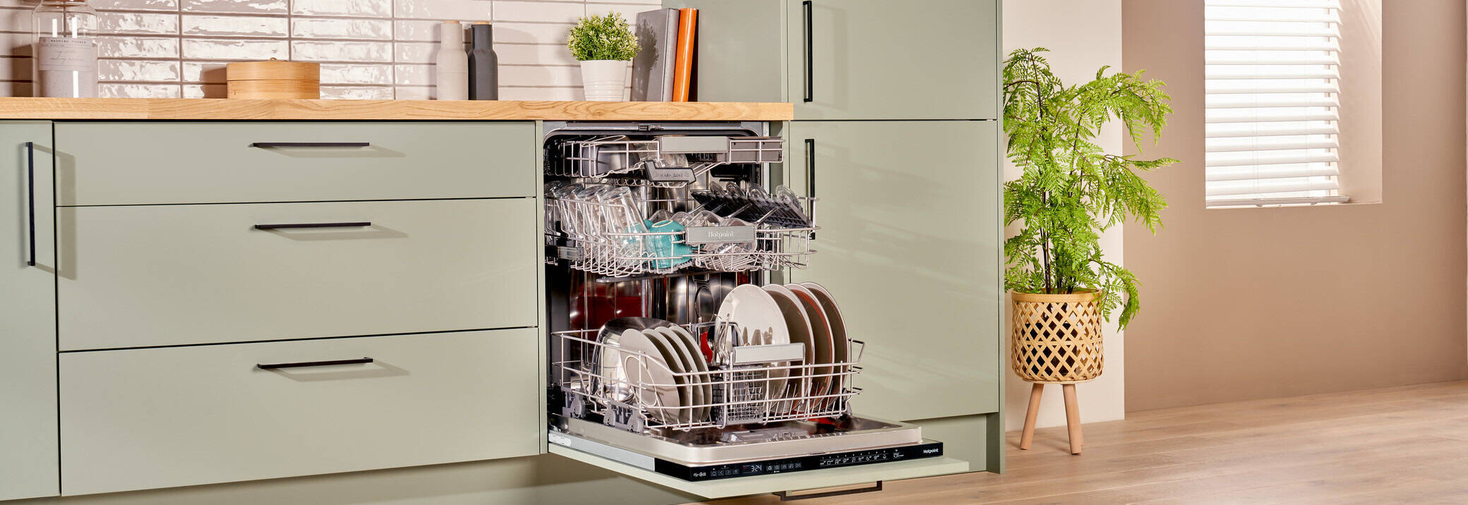 An open dishwasher in a modern kitchen with sage green cabinets and wooden countertops. The dishwasher is filled with neatly arranged plates, bowls, and cutlery. The countertop above features decorative items including plants, jars, and mugs
