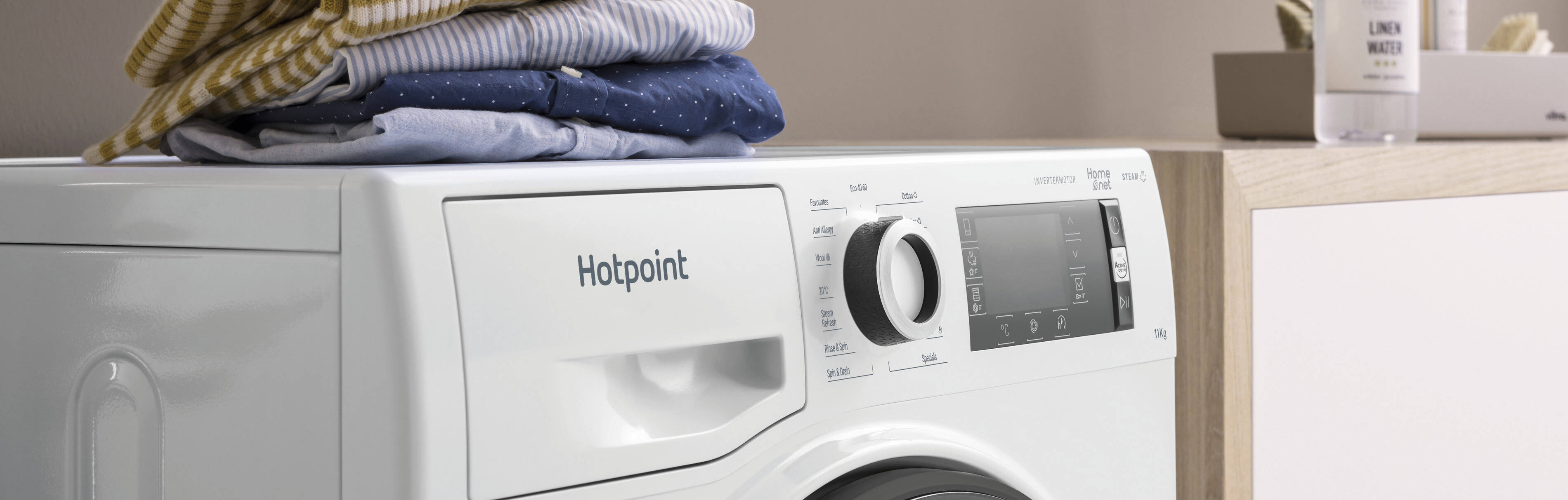 A sleek white Hotpoint washer dryer with a digital display sits on a wooden countertop with folded clothes and detergent bottles in a bright laundry room.