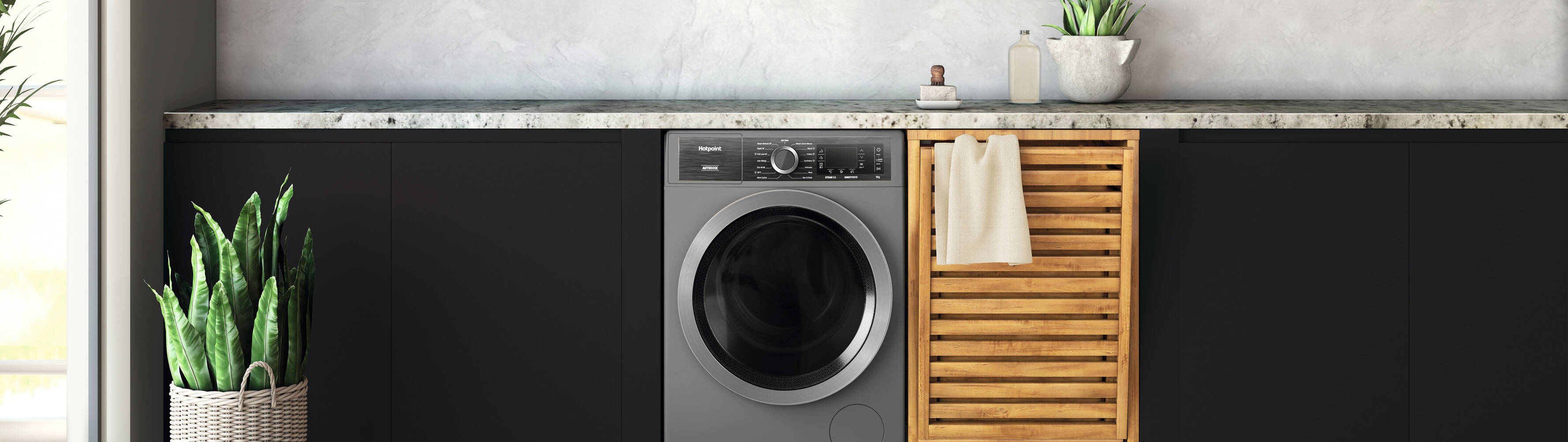 A bright laundry room with sleek black cabinets, a nice ceramic countertop, and a modern graphite washing machine, accented with green plants and a woven laundry basket on a light wood floor.