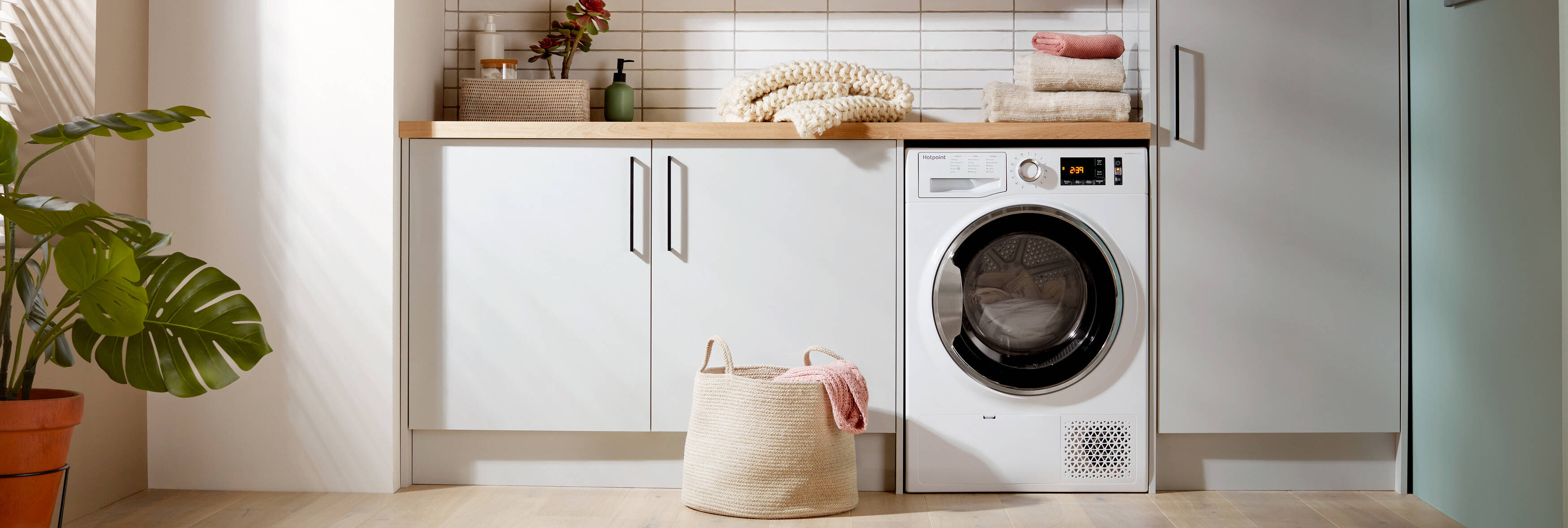 A laundry room with white cabinets, wooden countertops, and a tumble dryer. A laundry basket is on the floor, and folded towels sit on the counter near a potted plant.