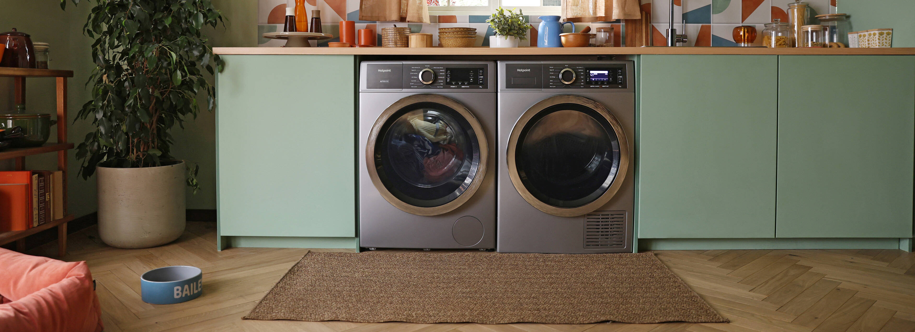 A laundry room with two front-loading washers under a wooden countertop. The room has green cabinets, colorful tile backsplash, a window, and a potted plant.