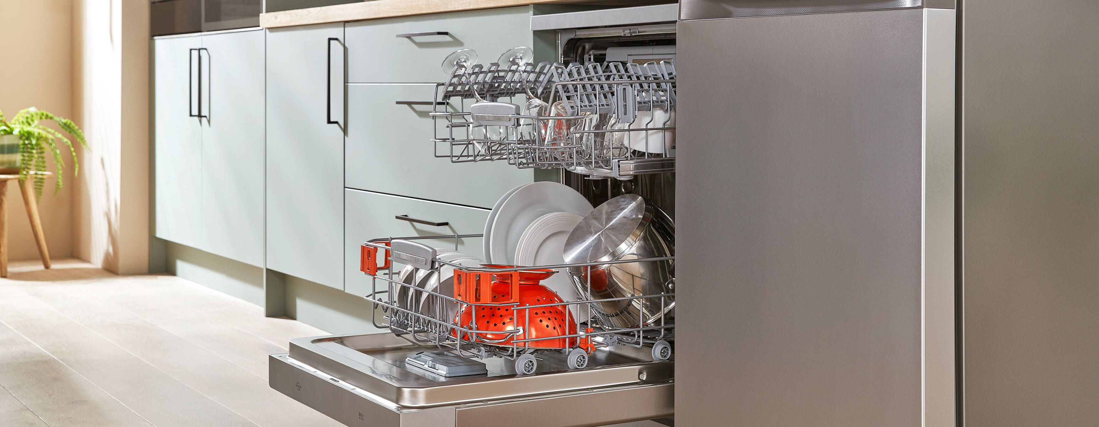 A man kneels in front of an open dishwasher, placing a saucepan into the bottom rack, which is partially loaded with plates and utensils.